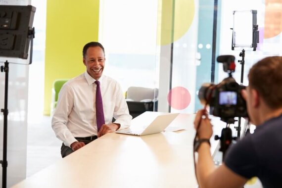 Executivo sentado em uma mesa montada em um cenário, sorrindo para a câmera, simbolizando o blogpost quando a sua marca precisa estar ao vivo nas redes sociais.