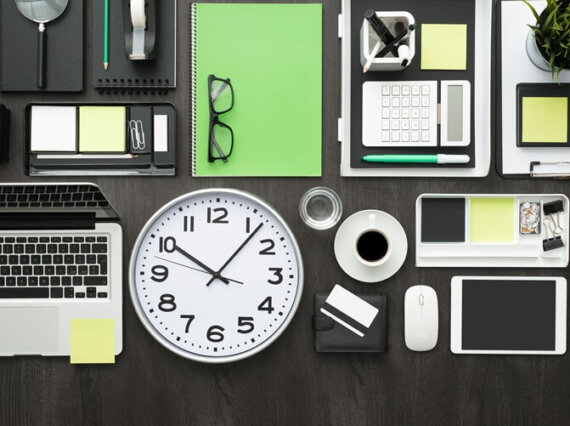 mesa com vários itens organizados: relogio computador, post-it, celular, óculos e agenda