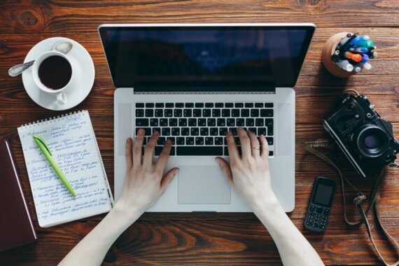 mulher digitando no notebook, sob uma mesa com bloco de anotações, uma xícara, celular e uma máquina fotográfica antiga