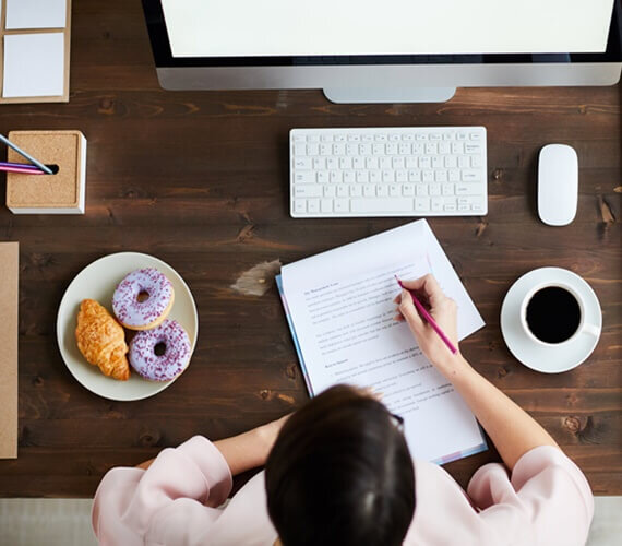 jovem sentada na frente de um computador fazendo anotações em um caderno, com um café e um donuts. simbolizando o blogpost sobre a necessidade ou não dos designers passarem pela faculdade.