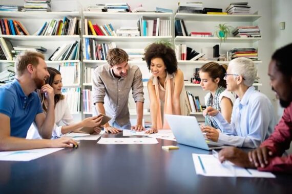 pessoas sentadas em uma mesa de reunião analisando papeis e computadores, com uma estante de livros no fundo