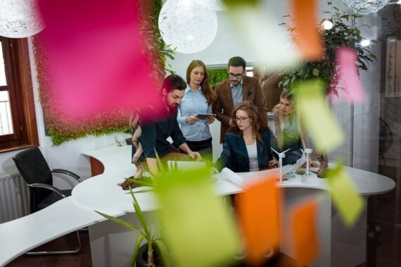 parede de vidro com post-it desfocados e ao fundo pessoas reunidas trabalhando