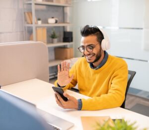 Homem sorrindo sentado em uma mesa de escritório, com celular na mão fazendo uma chamada de video