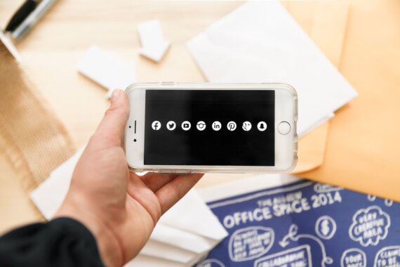 Mão segura celular e na tela vemos ícones de várias redes sociais, uma ilustração de um artigo sobre o slow content, que propõe desacelerar na produção e consumo de conteúdo online